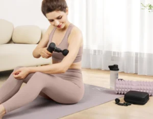 mujer sentada en el suelo vestida con unas mallas y top de color beig, dando uso a una mini pistola de masajes despues de hacer ejercicio