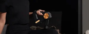 PISTOLA DE MASAJE THERAGUN COLOR NEGRO Y DORADO SIENDO UTILIZADA PÒR UNA MUJER VESTIDA DE NEGRO